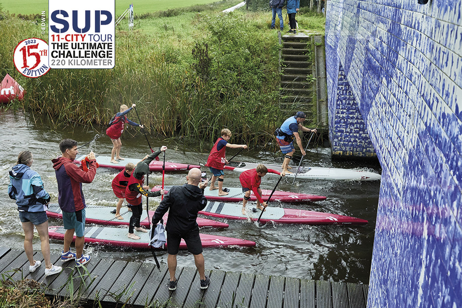 Part-of-the-event-ethos-,-encouraging-young-SUP-athletes,-here-passing-under-an-iconic-bridge-decorated-with-ice-skater-motifs-commemorating-the-origins-of-the-11Cities.